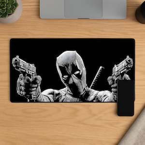 May include: Black rectangular mousepad featuring a black and white illustration of Deadpool holding two guns. The character is in a full-body pose, with a stern expression. A smartphone is visible on the right side of the mousepad.