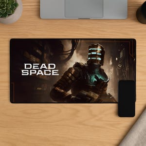 Può includere: Un tappetino per mouse rettangolare con la scritta "DEAD SPACE" in bianco. L'immagine raffigura un personaggio in tuta futuristica con un casco luminoso, su sfondo scuro e industriale. Uno smartphone nero è appoggiato a destra.