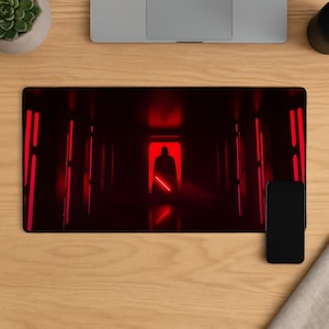 May include: A rectangular desk mat featuring a red-lit corridor with a silhouetted figure holding a lightsaber. The mat has a black border and is on a wooden desk. A black smartphone and a laptop are also visible.