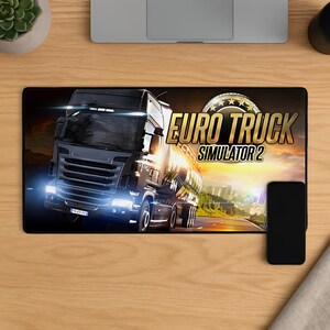 Op de afbeelding: Zwart en grijze muismat met een vrachtwagen en de tekst "Euro Truck Simulator 2" in goud. De achtergrond toont een zonsondergang boven een snelweg. Een zwarte smartphone ligt rechts op de muismat.