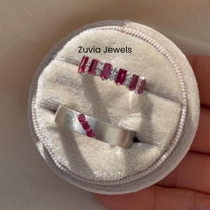 May include: Two rings displayed in a gray velvet jewelry box. One ring features a row of rectangular red gemstones and small white gemstones. The other ring is silver with a diagonal row of red gemstones. The text "Zuvia Jewels" is visible.