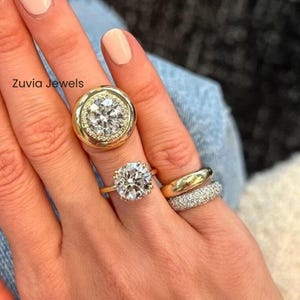 May include: A hand adorned with four gold rings. The rings showcase large, clear gemstones and diamond detailing. One ring features a large, round gemstone encircled by smaller diamonds. The rings are set against a denim background.