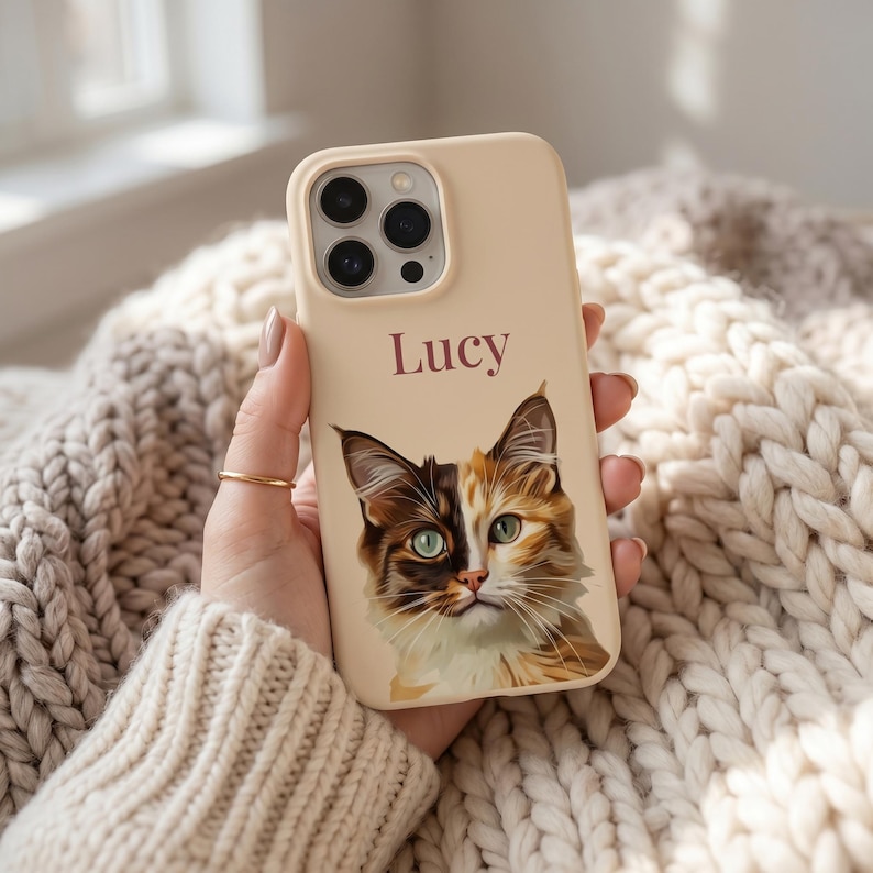 K&ouml;nnte beinhalten: Eine beige Smartphone-H&uuml;lle mit einer individuellen Illustration einer Gl&uuml;ckskatze mit gr&uuml;nen Augen und dem Namen Lucy in dunkelbrauner Schrift &uuml;ber der Katze. Die H&uuml;lle hat eine Aussparung f&uuml;r ein Dreifach-Kamerasystem oben links.