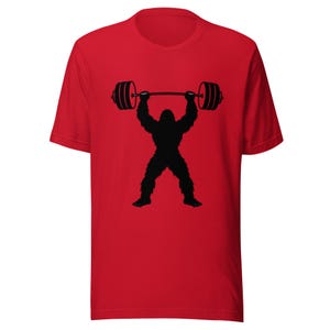 May include: Red t-shirt featuring a black silhouette of a Bigfoot lifting a barbell overhead. The design showcases a weightlifting theme, perfect for fitness enthusiasts. The shirt is a classic crew neck style.