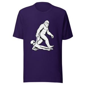 May include: A purple t-shirt featuring a black and white graphic of Bigfoot holding an alien. The Bigfoot is walking, and the alien is lying on the ground. The shirt is made of a soft material.