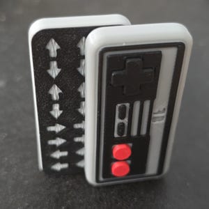 May include: Two rectangular gaming controller magnets. One features a grey border with a black control panel, a black directional pad, two small black buttons, and two round red buttons. The other is black with a white arrow pattern.