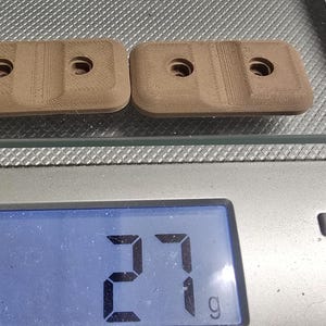 May include: Two rectangular, light brown, 3D-printed plastic components with rounded corners and two circular holes each, resting on a digital kitchen scale that displays a weight of 27 grams, indicating the combined mass of the two items.