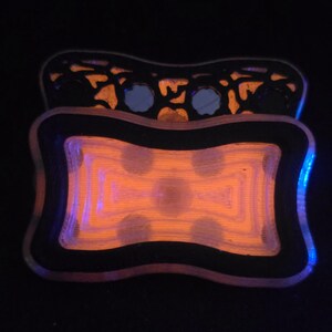 May include: A rectangular tray with rounded corners featuring a black border and a glowing orange center with a concentric geometric pattern. A second, similar tray with a black filigree cutout design is positioned directly behind the first tray.