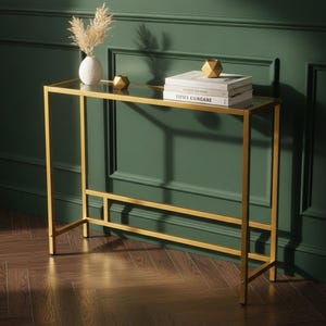 May include: A rectangular console table with a gold-toned metal frame and a clear glass top. On the table sits a white vase with dried pampas grass, a small gold geometric object, and two stacked books with the text Vogue Living and Time Curated.