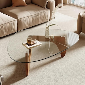 May include: A modern, oval-shaped coffee table with a clear glass top and wooden legs. The table is in a living room setting, with a beige sofa and chair visible. A small table with a drink and book is on the table.