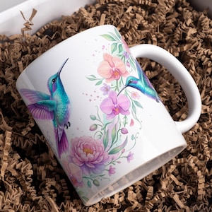 May include: White ceramic mug featuring a vibrant design of two hummingbirds with turquoise and purple plumage, surrounded by pink and purple flowers and green leaves. The mug has a white handle and is presented in a box with brown paper filler.