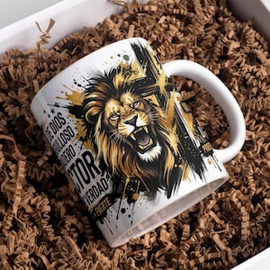May include: A white ceramic mug featuring a detailed graphic illustration of a roaring lion in shades of gold, black, and white. The design includes abstract paint splatters and Spanish text including the words DE DIOS, ORGULLOSO, VERDAD, and FUERTE.