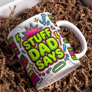 Op de afbeelding: Witte keramische mok met de tekst "STUFF DAD SAYS" in grote groene letters, omringd door kleurrijke graphics en zinnen. De mok heeft een wit oor en wordt gepresenteerd in een geschenkdoos met bruin papier.