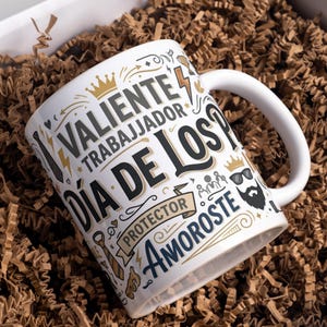 May include: White ceramic mug with a handle, featuring the text "VALIENTE TRABAJADOR DÍA DE LOS PROTECTOR AMOROSTE" in black, gold, and gray. The mug is decorated with crowns, lightning bolts, and other graphic elements. The mug is presented in a box with brown paper.