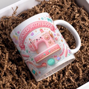 Pode incluir: Uma caneca de cerâmica branca com uma ilustração de uma máquina de costura rosa, o texto "CREATED WITH LOVE" e vários gráficos relacionados à costura, incluindo uma fita métrica, carretéis de linha e um novelo de lã em tons pastel.