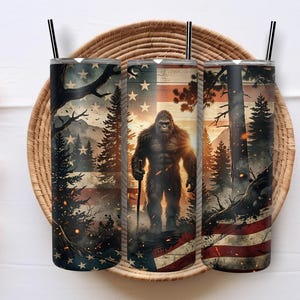Pode incluir: Três copos cilíndricos com palhinhas pretas com uma impressão gráfica de uma criatura Bigfoot numa floresta com uma bandeira americana ao fundo. O design inclui pinheiros, montanhas e brasas brilhantes em vermelho, branco e azul.