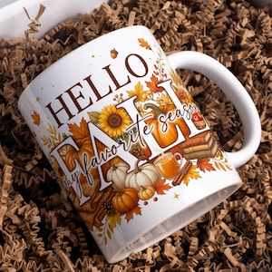 Op de afbeelding: Een witte keramische mok met de tekst HELLO FALL my favorite season in bruine en witte letters. Het ontwerp bevat illustraties van oranje pompoenen, gele zonnebloemen, herfstbladeren, een stapel boeken en een warme drank.