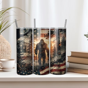 May include: A cylindrical metal tumbler featuring a graphic print of a Bigfoot creature standing in a forest with a sunset background and an American flag motif. The design includes dark pine trees, glowing embers, and a silver straw inserted at the top.