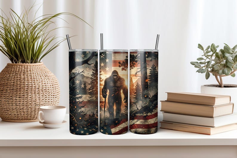 May include: A cylindrical metal tumbler featuring a graphic print of a Bigfoot creature standing in a forest with a sunset background and an American flag motif. The design includes dark pine trees, glowing embers, and a silver straw inserted at the top.