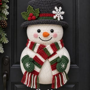May include: A crocheted snowman door hanger featuring a dark gray top hat with holly and a white snowflake, a red, white, and green striped scarf, and green mittens with white snowflake embroidery. The text reads Jolly THE SNOWMAN FESTIVE DOOR HANGER.
