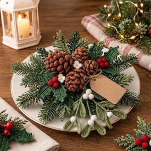 May include: A decorative arrangement featuring crocheted pinecones, green holly leaves, red berries, and white mistletoe berries on a white plate. A blank brown paper tag is tied to the arrangement with twine, creating a festive holiday centerpiece.