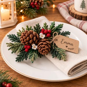 May include: A beige linen napkin featuring a crocheted holiday decoration with two brown pinecones, green cedar sprigs, small red berries, and white flowers. A rectangular kraft paper gift tag is attached, printed with the name Emma and a heart.