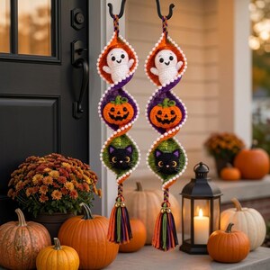May include: Two vertical crochet spiral decorations featuring a white ghost, an orange jack-o'-lantern, and a black cat face in each section. The spirals are edged with small white pom-poms and finished with multi-colored yarn tassels at the bottom.