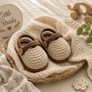 May include: A pair of cream and brown crocheted baby shoes with a strap and a small wooden button on each side. The shoes are placed in a woven basket alongside a cream crocheted blanket, a wooden elephant toy, and a round sign reading Little Strides.