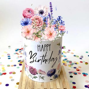 May include: A white cylinder-shaped birthday cake decorated with a floral arrangement of pink, blue, and purple flowers. The words "Happy Birthday" are written in black script on the front of the cake. The cake sits on a wooden surface, with colorful confetti scattered around.