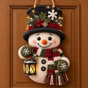 May include: A crocheted snowman wall hanging featuring a black top hat adorned with holly, berries, and a white snowflake. The snowman wears a red, white, and green striped scarf, has two black button accents, and holds a small lit lantern.