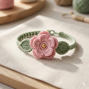 Op de afbeelding: Een gehaakte armband met een centrale roze bloem met vijf bloemblaadjes en een kleine goudkleurige kraal in het midden. De band is gemaakt van saliegroen garen met twee gehaakte bladvormen aan weerszijden van de bloem voor een bloemmotief.