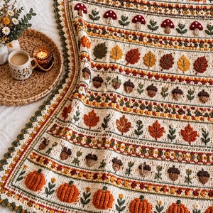 May include: A cream-colored crochet blanket featuring horizontal rows of textured autumn motifs including orange pumpkins, red mushrooms with white spots, brown acorns, and various fall leaves in shades of yellow, orange, and green.