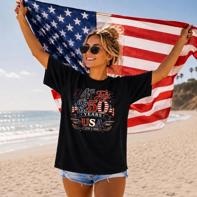 May include: A black short-sleeved t-shirt featuring a graphic print with the text 4th July 250 Years USA 1776-2026, decorated with stars, stripes, and fireworks motifs. The shirt is worn with light blue denim shorts and dark sunglasses.