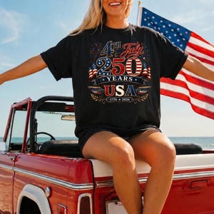 May include: A black short-sleeve t-shirt featuring a graphic design with the text 4th July 250 Years USA 1776-2026. The design includes American flag patterns, fireworks, and decorative flourishes in red, white, and blue colors on the front of the shirt.