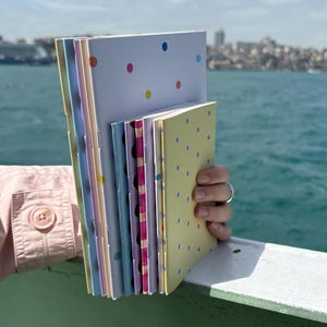 May include: A stack of colorful notebooks with polka dot patterns in various colors, including pink, blue, and yellow. The notebooks are held in front of a blurred background of water and a city skyline.