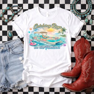 May include: A white t-shirt featuring a graphic print of a person relaxing on a pontoon boat in the ocean at sunset. The design includes palm trees, a pink flamingo float, seashells, and the text Catching Rays on the Pontoon in blue and gold.