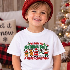 May include: A white short-sleeved t-shirt featuring a colorful graphic of five Christmas characters facing away with their backsides showing. The text reads: WE WISH YOU NOTHING BUTT A MERRY CHRISTMAS. The characters include a gingerbread man, reindeer, Santa, snowman, and elf.