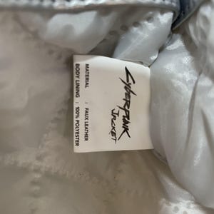 May include: Close-up of a tag from a jacket. The tag reads "Cyberpunk Jacket" in a stylized font. The material is faux leather, and the body lining is 100% polyester. The jacket appears to be light blue.