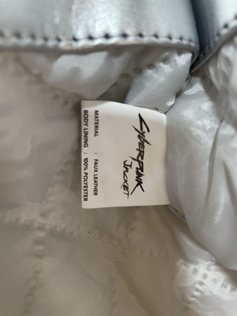 May include: Close-up of a tag from a jacket. The tag reads "Cyberpunk Jacket" in a stylized font. The material is faux leather, and the body lining is 100% polyester. The jacket appears to be light blue.