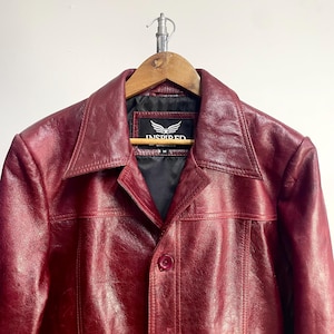 May include: A deep red leather jacket with a pointed collar, a single front button closure, and horizontal seam detailing across the chest. The interior label features the text INSPIRED JACKETS with a winged logo and the size M printed below.