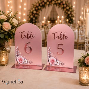 May include: Two arched rose gold table number signs labeled Table 6 and Table 5. Each sign features a smooth metallic finish with a floral illustration of pink roses and white flowers at the bottom corner. The numbers are in a bold, dark font.