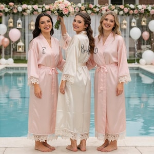 Può includere: Tre donne con vestaglie di raso. Due sono rosa chiaro con finiture in pizzo, e una è avorio con "Bride" stampato sul retro. Le vestaglie hanno una cintura annodata e arrivano al ginocchio. Una vestaglia rosa ha la lettera "C" sul davanti.