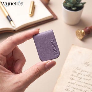May include: A small, rectangular, lavender-colored magnetic bookmark with a textured surface. The word MRSFIN is embossed vertically in a clean, sans-serif font on the front of the bookmark. The item has rounded corners and a folded design.
