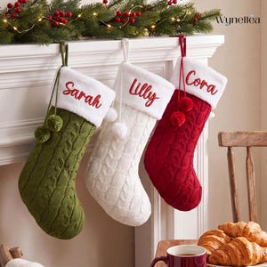 Può includere: Tre calze natalizie in maglia a trecce nei colori verde oliva, bianco e rosso. Ogni calza presenta un risvolto in peluche bianco ricamato con i nomi Sarah, Lilly e Cora in corsivo rosso. Due pon-pon abbinati pendono dalla parte superiore.