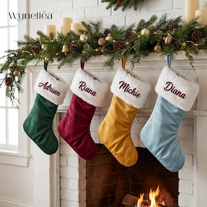 Può includere: Quattro calze natalizie in velluto con risvolti in pelliccia sintetica bianca, ognuna ricamata con un nome: Adrian in verde, Riana in bordeaux, Michie in oro e Diana in azzurro. Le calze sono appese in fila e presentano una texture morbida.