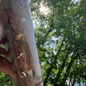 May include: A gold-colored chain bookmark with various charms, including a pearl, a heart, and a small key, is draped over a Bible. The Bible has a marbled cover with the words "Holy Bible" embossed on it. The background features lush green trees and sunlight.