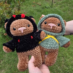 May include: Two handmade crochet teddy bears. One bear wears a black hoodie with red horns, the other a green hoodie with a yellow sash. Both have brown bodies, pink noses, and small fangs. The bears are plush and soft.