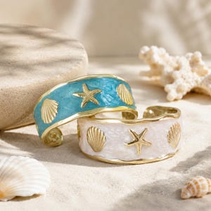 Peut inclure: Deux bracelets ouverts de type manchette au thème côtier. L'un présente une finition en émail bleu clair, l'autre une finition en émail blanc. Les deux bracelets sont ornés de motifs d'étoiles de mer et de coquillages en métal doré.