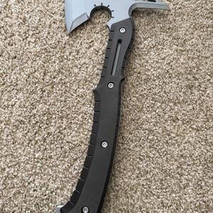May include: A tactical multi-tool hatchet featuring a silver-toned metal axe head with a curved blade and a pointed rear spike. The handle is constructed from black textured material, secured with silver screws, and includes a lanyard hole at the base.