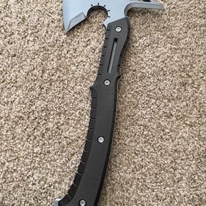 May include: A multi-functional tool featuring a silver-colored axe head with a curved blade and a pointed rear spike. The handle is black with a textured grip, secured by three visible silver screws, and includes a lanyard hole at the base.